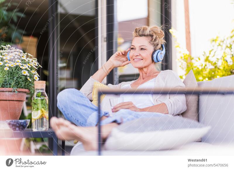 Porträt einer lächelnden reifen Frau, die auf der Terrasse sitzt und mit Kopfhörern Musik hört Flaschen Glasflaschen Gläser Trinkglas Trinkgläser Couch Couches