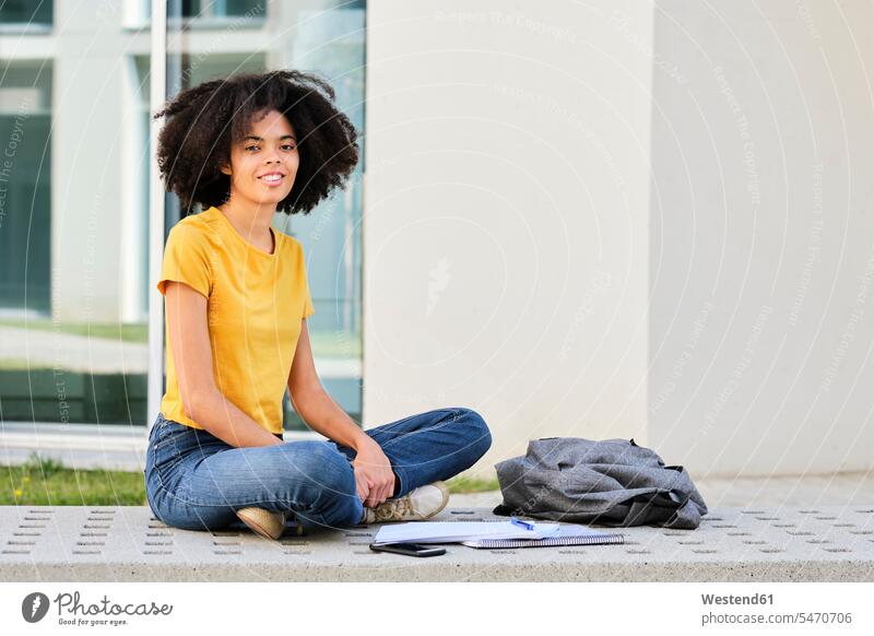 Junge Studentin sitzt mit gekreuzten Beinen durch Bücher und Tasche auf dem Campus Farbaufnahme Farbe Farbfoto Farbphoto Außenaufnahme außen draußen im Freien