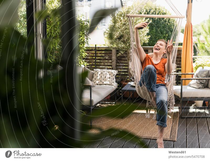 Fröhliche Frau, die sich mit einem Smartphone auf einer Schaukel auf der Veranda sitzend durch die Tür sieht Farbaufnahme Farbe Farbfoto Farbphoto Deutschland