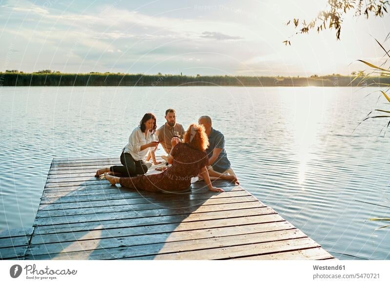 Freunde beim Picknick auf einem Steg an einem See bei Sonnenuntergang Leute Menschen People Person Personen Europäisch Kaukasier kaukasisch Gruppe von Menschen