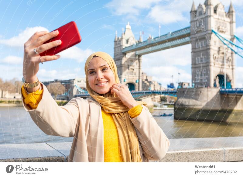 Porträt einer lächelnden jungen Frau, die ein Selfie mit ihrem Smartphone vor der Tower Bridge macht, London, UK Leute Menschen People Person Personen