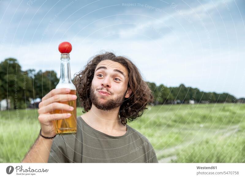 Porträt eines lächelnden jungen Mannes im Freien, der eine Tomate auf einer Bierflasche balanciert Bierflaschen balancieren Balance Portrait Porträts Portraits