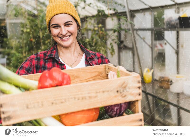 Lächelnde Frau, die eine Kiste mit Gemüse trägt, während sie vor einem Gewächshaus steht Farbaufnahme Farbe Farbfoto Farbphoto Deutschland Freizeitkleidung