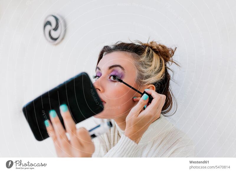 Junge Frau trägt Wimperntusche auf, während sie mit einem Smartphone vor einer weißen Wand steht Farbaufnahme Farbe Farbfoto Farbphoto Außenaufnahme außen