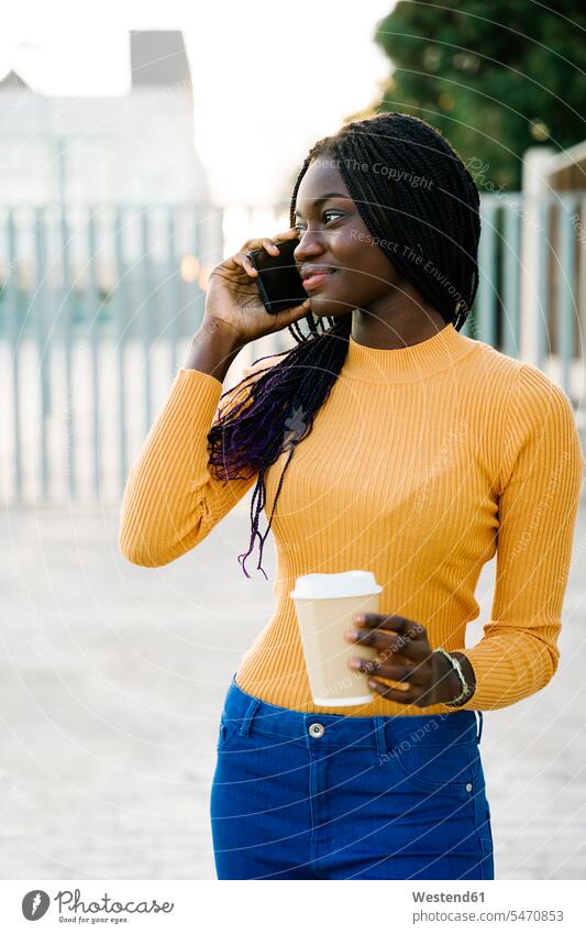 Lächelnde Teenager-Mädchen mit Kaffeetasse Blick weg während des Gesprächs auf Smartphone auf der Straße Farbaufnahme Farbe Farbfoto Farbphoto Außenaufnahme