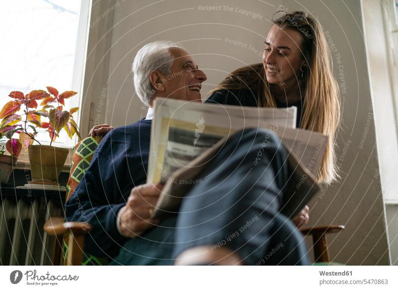Glückliche junge Frau und älterer Mann mit Zeitung zu Hause Generation Zeitungen Stuehle Stühle Lektüre sitzend sitzt freuen Frohsinn Fröhlichkeit Heiterkeit