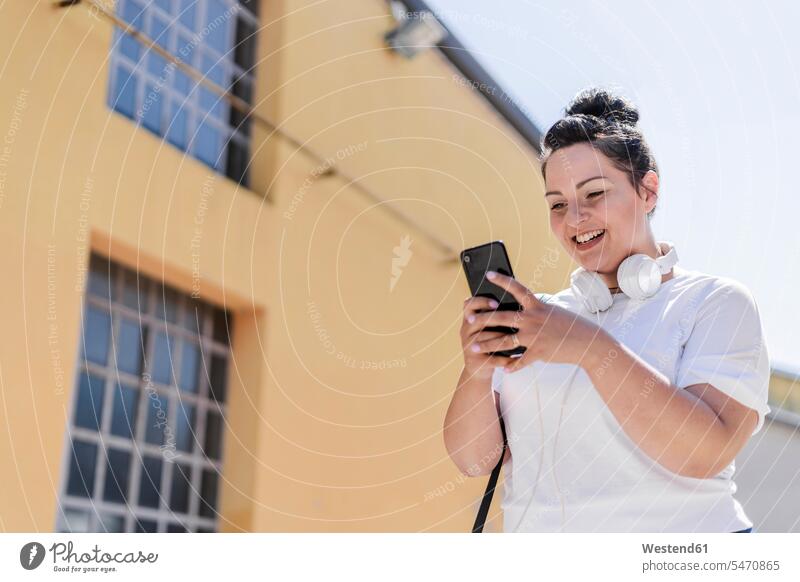 Fröhliche kurvige junge Frau mit Kopfhörern, die in der Stadt mit dem Handy telefoniert Leute Menschen People Person Personen Europäisch Kaukasier kaukasisch 1