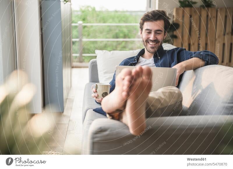 Junger Mann sitzt auf Couch, benutzt Laptop, trinkt Kaffee Deutschland Laptop benutzen Laptop benützen offenes Lächeln lachen offenes Laecheln
