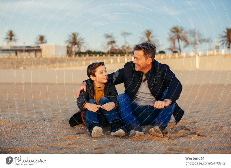 Lächelnder Vater umarmt Sohn am Strand Beach Straende Strände Beaches lächeln sitzen sitzend sitzt Söhne umarmen Umarmung Umarmungen Arm umlegen Papas Väter