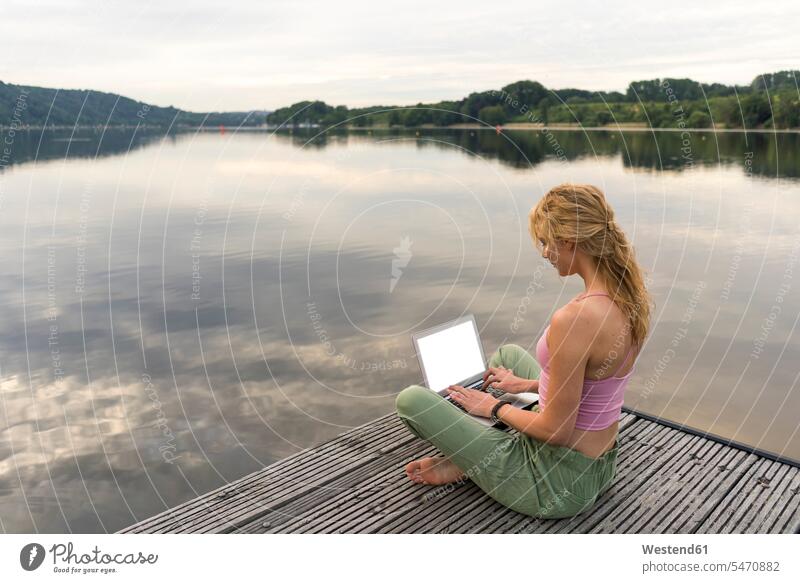 Junge Frau mit Laptop auf einem Steg an einem See Job Berufe Berufstätigkeit Beschäftigung Jobs Rechner Laptops Notebook Notebooks Arbeit sitzend sitzt früh
