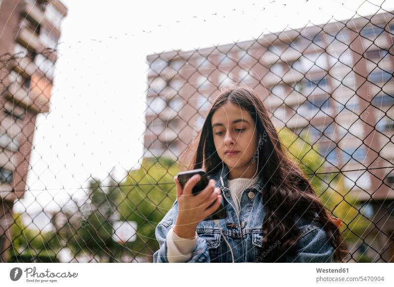 Niedriger Blickwinkel auf ein Teenager-Mädchen, das ein Smartphone benutzt, während es gegen einen Kettenzaun steht Farbaufnahme Farbe Farbfoto Farbphoto