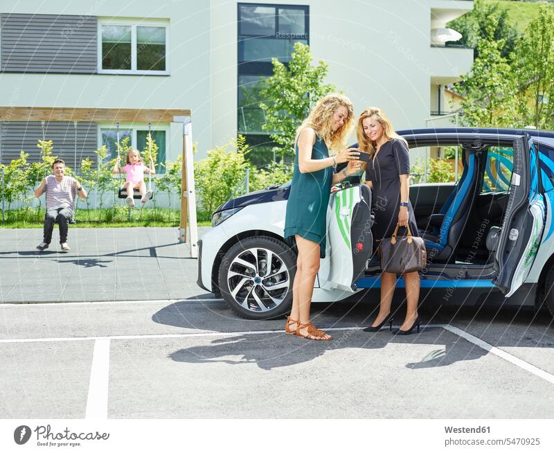 Zwei Frauen mit Handy am Elektroauto und Mann und Mädchen im Hintergrund Elektromobil Elektromobile Elektroautos Auto Wagen PKWs Automobil Autos weiblich