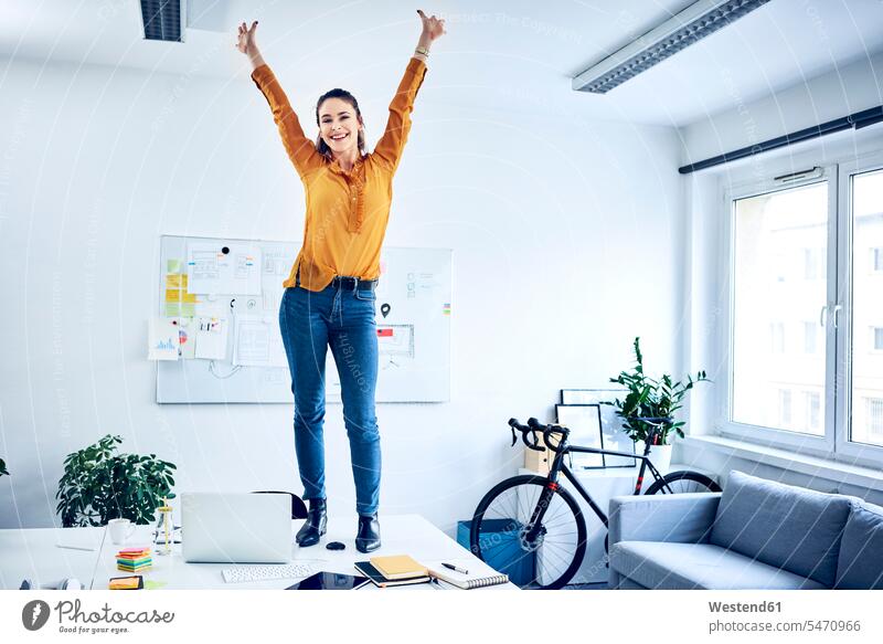 Glückliche junge Geschäftsfrau jubelnd im Büro stehend auf dem Schreibtisch glücklich glücklich sein glücklichsein steht Jubel Office Büros Arbeitstisch
