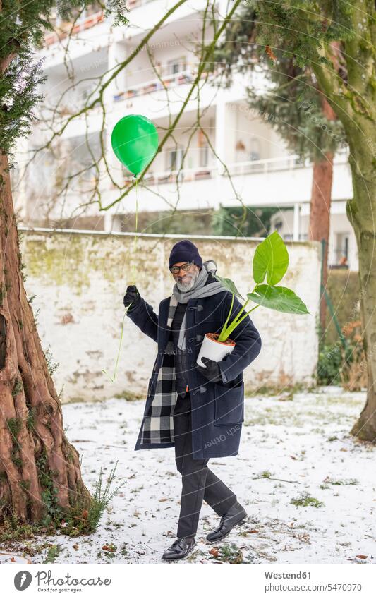 Lächelnder reifer Mann mit grünem Ballon und Topfpflanze im Winter lächeln Luftballon Luftballons Ballons Luftballone Männer männlich winterlich Winterzeit