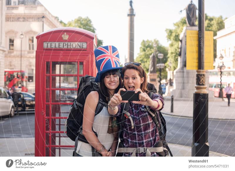 Fröhliche Frau, die ein Selfie mit einem Freund macht, der einen Hut mit britischer Flagge vor einer roten Telefonzelle in der Stadt trägt Farbaufnahme Farbe