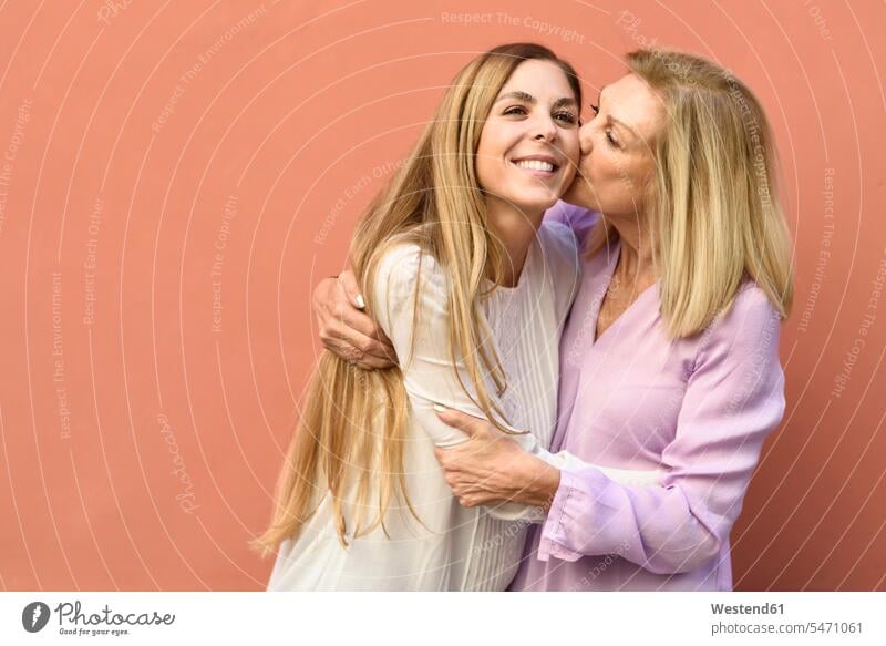 Glückliche reife Frau umarmt und küsst ihre Tochter an einer roten Wand Freunde Kameradschaft Freundin Kuss Küsse Arm umlegen Umarmung Umarmungen freuen