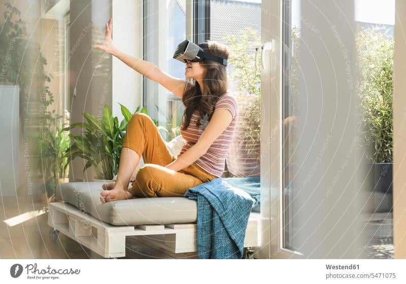 Junge Frau sitzt am Fenster zu Hause mit VR-Brille virtuell Virtualität weiblich Frauen Zuhause daheim sitzen sitzend Brillen Erwachsener erwachsen Mensch