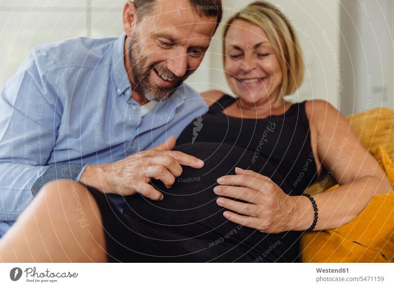 Älterer Mann und seine schwangere reife Frau sitzen auf der Couch und berühren ihren Bauch sitzend sitzt Bäuche Berührung anfassen Schwangere schwangere Frau