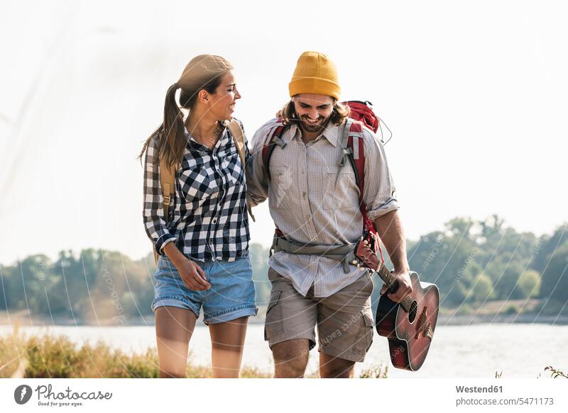 Glückliches junges Paar mit Rucksäcken und Gitarre beim Spaziergang am Flussufer Rucksack glücklich glücklich sein glücklichsein Fluesse Fluß Flüsse Pärchen