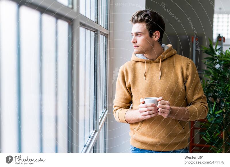 Junger Mann mit Kaffeetasse nachdenklich beim Blick durch Fenster in neue Wohnung Farbaufnahme Farbe Farbfoto Farbphoto Innenaufnahme Innenaufnahmen innen