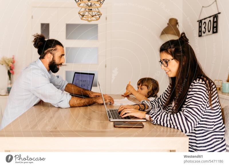 Frau arbeitet am Laptop, während Vater und Tochter zu Hause am Esstisch malen Farbaufnahme Farbe Farbfoto Farbphoto Innenaufnahme Innenaufnahmen innen drinnen