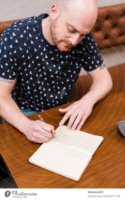 Mittelgroßer erwachsener Mann konzentriert sich beim Schreiben in einem Notizbuch im Café sitzend Farbaufnahme Farbe Farbfoto Farbphoto Innenaufnahme