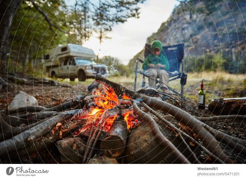 Chile, Santa Magda, Rio Maniguales, Junge sitzt am Lagerfeuer im Wald mit Wohnmobil im Hintergrund Buben Knabe Jungen Knaben männlich sitzen sitzend Forst