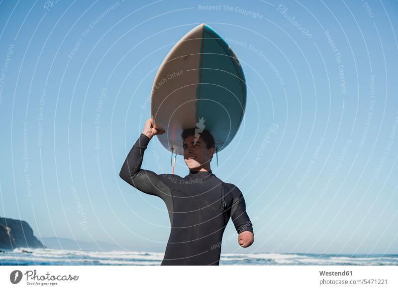 Behinderter Surfer, der sein Surfbrett auf dem Kopf trägt Surfing Wellenreiten surfboard surfboards Surfbretter freuen Glück glücklich sein glücklichsein