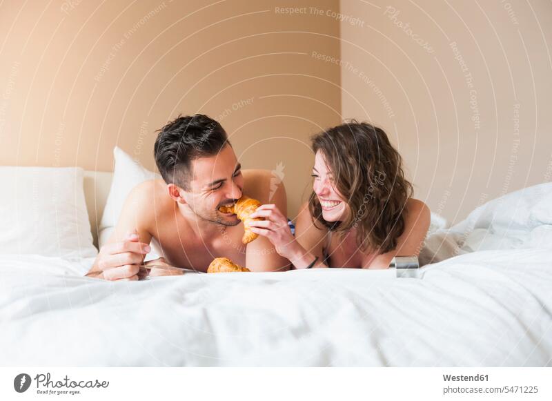 Glückliche junge Frau füttert Mann mit Croissant, während sie zu Hause im Bett liegt Farbaufnahme Farbe Farbfoto Farbphoto Innenaufnahme Innenaufnahmen innen