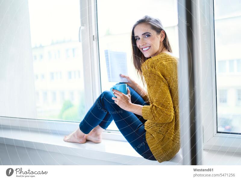 Junge Frau sitzt auf der Fensterbank mit Buch und Tasse Tee und schaut in die Kamera Leute Menschen People Person Personen Alleinstehende Alleinstehender