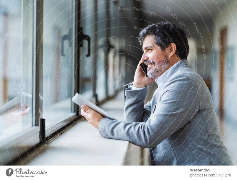 Lächelnder männlicher Professor telefoniert, während er durch ein Fenster im Korridor der Universität schaut Farbaufnahme Farbe Farbfoto Farbphoto Tag