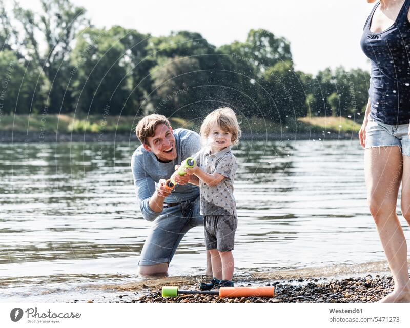 Vater und Sohn haben Spaß am Flussufer, spielen mit einer Wasserpistole Wasserkanone Wasserspritze Spass Späße spassig Spässe spaßig lachen Papas Väter Vati