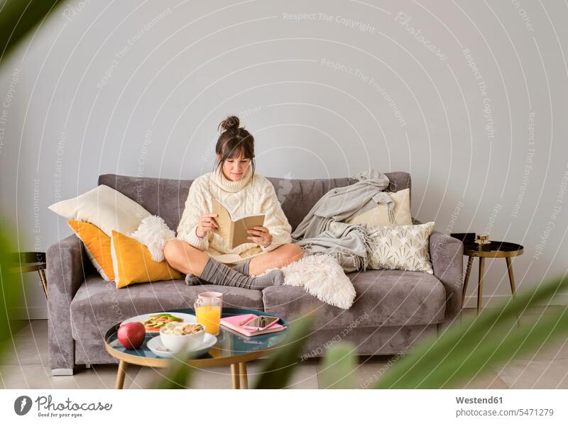 Frau, die ein Buch liest, während sie auf dem Sofa gegen die Wand zu Hause im Winter sitzt Farbaufnahme Farbe Farbfoto Farbphoto Spanien Innenaufnahme