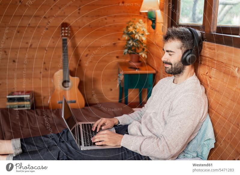 Lächelnder junger Mann, der einen Laptop benutzt, während er auf einem Bett in einer Blockhütte Musik hört Farbaufnahme Farbe Farbfoto Farbphoto Außenaufnahme