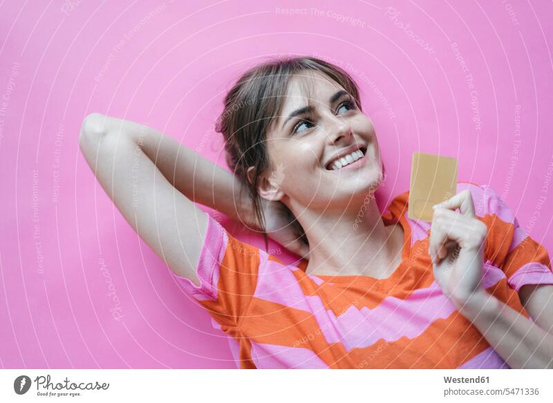 Auf rosa Hintergrund liegende Frau mit Händen hinter dem Kopf, die eine Kreditkarte halten EC-Karte Kreditkarten pink pinkfarben Hand hinter dem Kopf