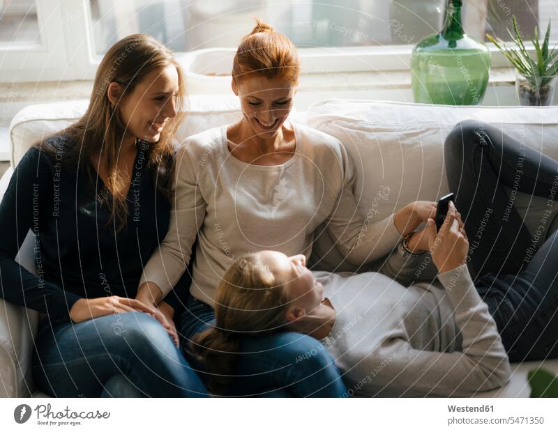 Glückliche Mutter mit zwei Teenager-Mädchen auf der Couch zu Hause mit Handy Teenagerin junges Mädchen Teenagerinnen weiblich junge Frau Zuhause daheim Sofa