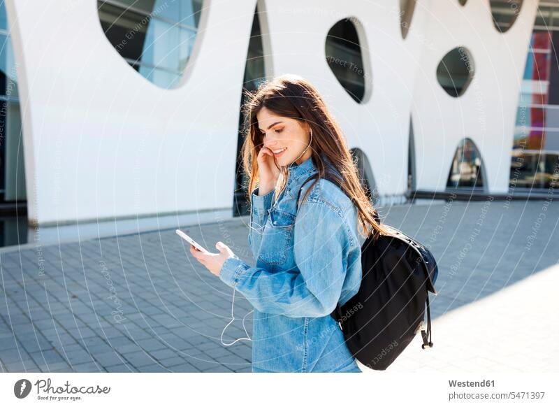 Spanien, Barcelona, lächelnde junge Frau mit Rucksack, die mit Handy und Kopfhörern Musik hört weiblich Frauen Ohrhörer Smartphone iPhone Smartphones Rucksäcke