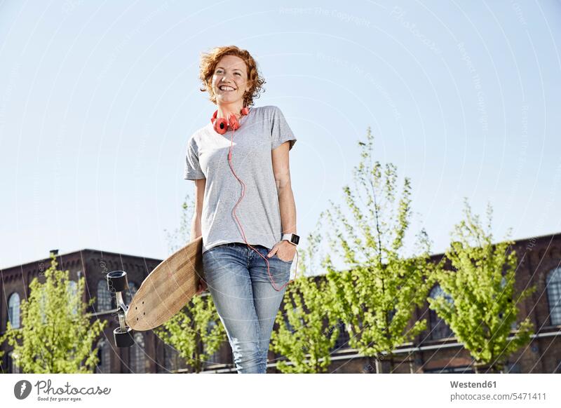 Lächelnde junge Frau mit Longboard, Kopfhörer und Smartwatch steht in städtischer Umgebung lächeln Kopfhoerer stehen stehend weiblich Frauen Urban Urbanität