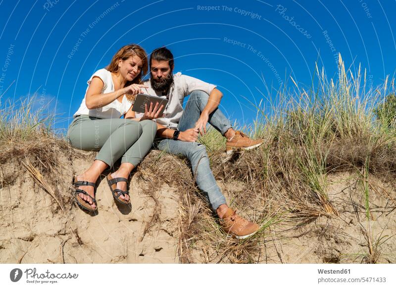Junges Paar sitzt auf einer Düne im Sommer, mit digitalem Tablet erholen erholend Junge Paare Sommerzeit sommerlich sitzen sitzend Duenen Dünen Tablet Computer