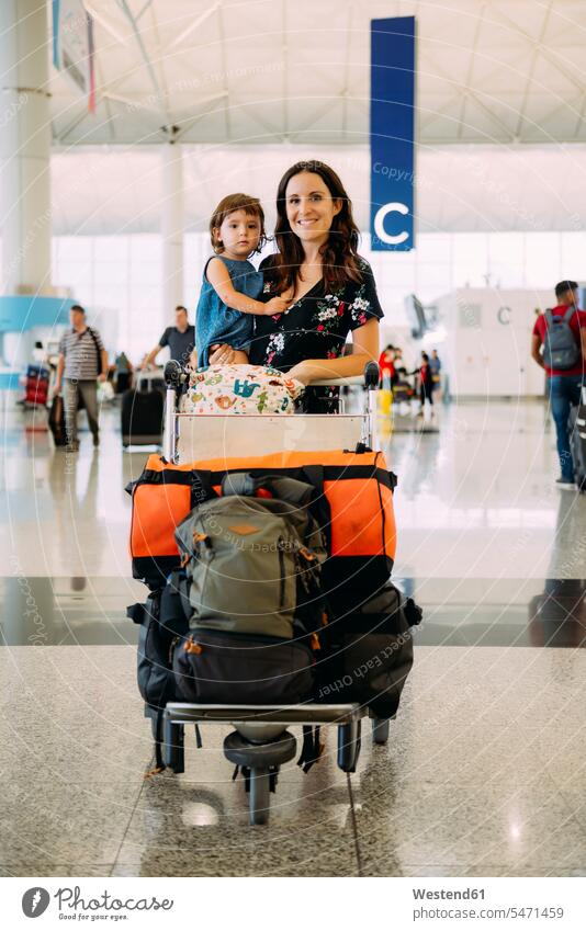 Mutter hält ein kleines Mädchen am Flughafen und schiebt einen Trolley mit dem Gepäck stehen stehend steht schieben anschieben lächeln Tochter Töchter