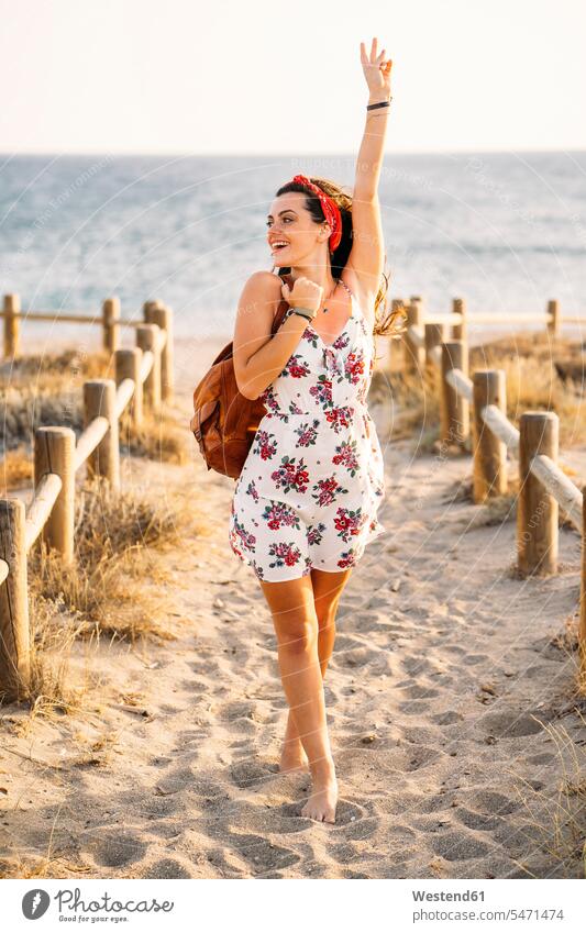 Junge Frau mit erhobenem Arm, die abends am Strand spazieren geht Zäune Rucksäcke gehend sommerlich Sommerzeit freuen Glück glücklich sein glücklichsein