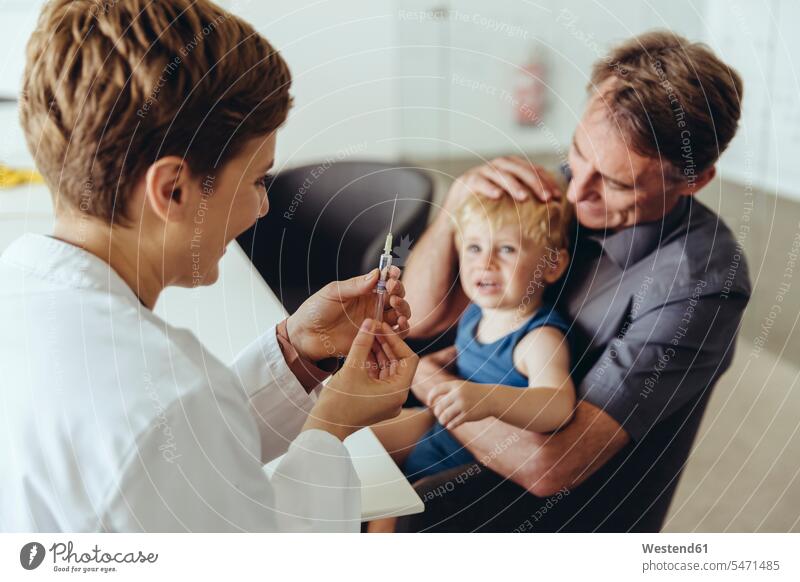 Vater hält seinen Sohn, während er von einem Kinderarzt geimpft wird Söhne Junge männliche Babys männliches Baby männliche Babies Papas Väter Vati Vatis Papis