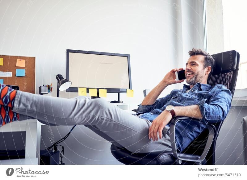Entspannter Mann sitzt am Schreibtisch im Büro und telefoniert mit dem Handy telefonieren anrufen Anruf telephonieren Office Büros Mobiltelefon Handies Handys