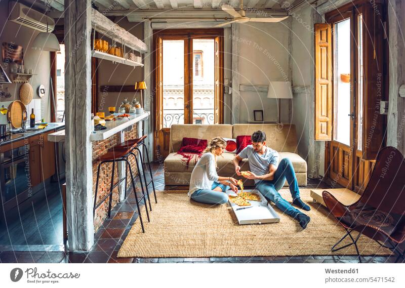 Junges Paar sitzt auf dem Boden des Wohnzimmers und isst Pizza aus dem Karton Boxen Kiste Schachtel Couch Couches Liege Sofas Teppiche sitzend daheim zu Hause