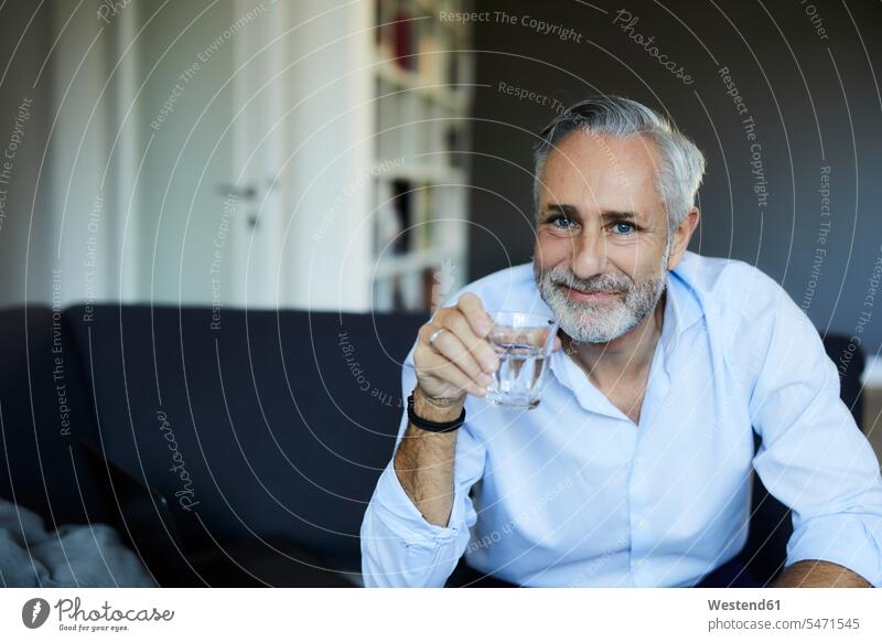Lächelnder reifer Mann, der zu Hause auf dem Sofa ein Glas Wasser trinkt Leute Menschen People Person Personen Alleinstehende Alleinstehender Singles