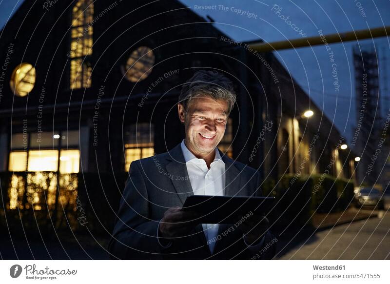 Lächelnder Geschäftsmann mit Tablette vor modernem Gebäude in der Nacht nachts lächeln Tablet Computer Tablet-PC Tablet PC iPad Tablet-Computer Businessmann