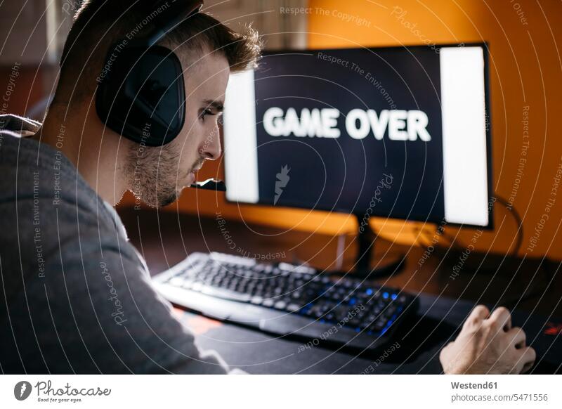Junger Mann sitzt an seinem PC und schaut auf den Bildschirm mit Game-Over-Text Kopfhörer Kopfhoerer Spannung Anspannung Verspannung Computerspiel