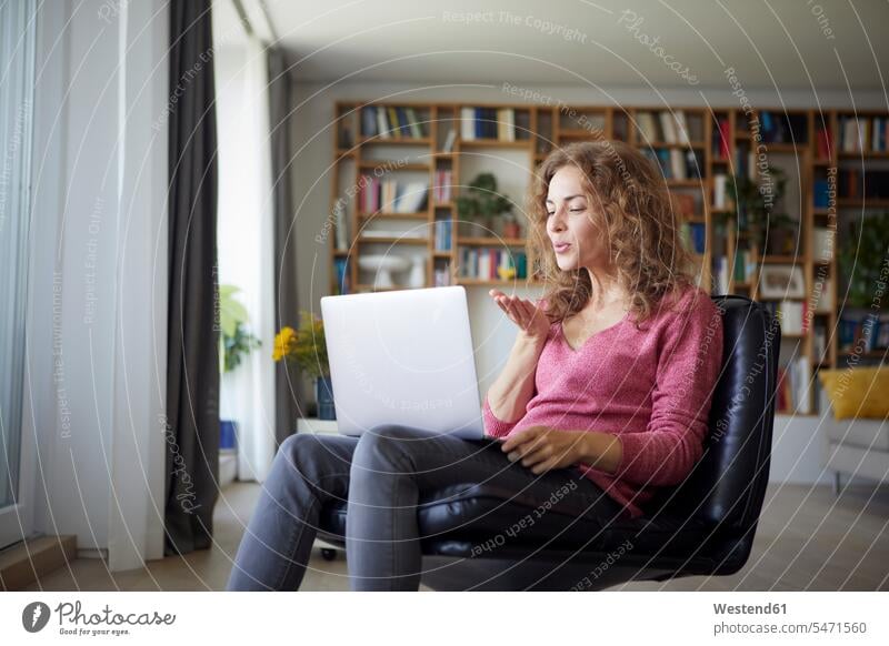 Frau bläst Kuss zu Videoanruf auf Laptop, während sie zu Hause sitzt Farbaufnahme Farbe Farbfoto Farbphoto Innenaufnahme Innenaufnahmen innen drinnen Tag