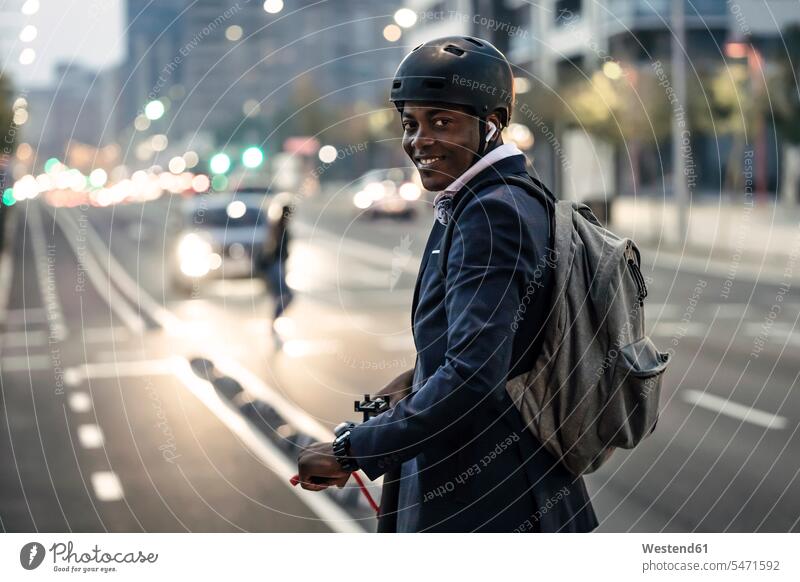 Porträt eines lächelnden Geschäftsmannes mit Schiebemotorroller auf dem Fahrradweg am Abend geschäftlich Geschäftsleben Geschäftswelt Geschäftsperson