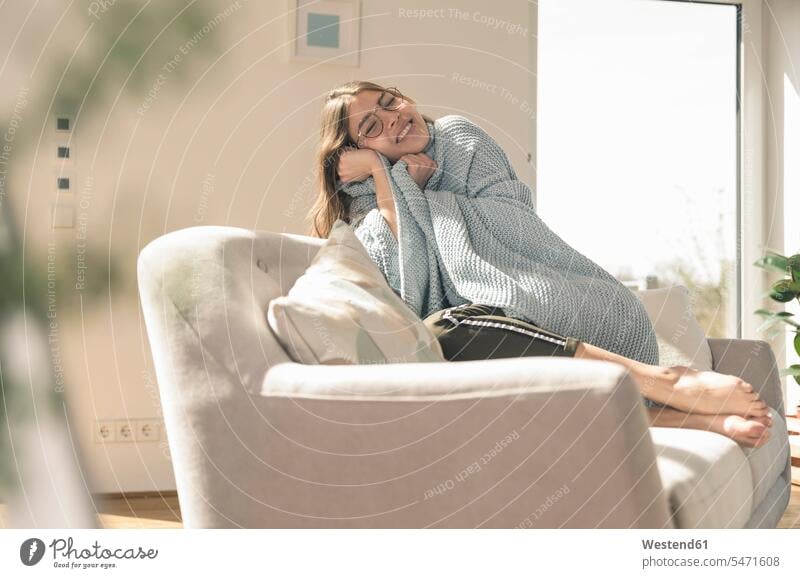 Glückliche junge Frau mit Wolldecke auf Couch Deutschland Wohnung wohnen Wohnungen Wohlbefinden Lifestyle Lebensstil Komfortabel Gemütlich Bequem